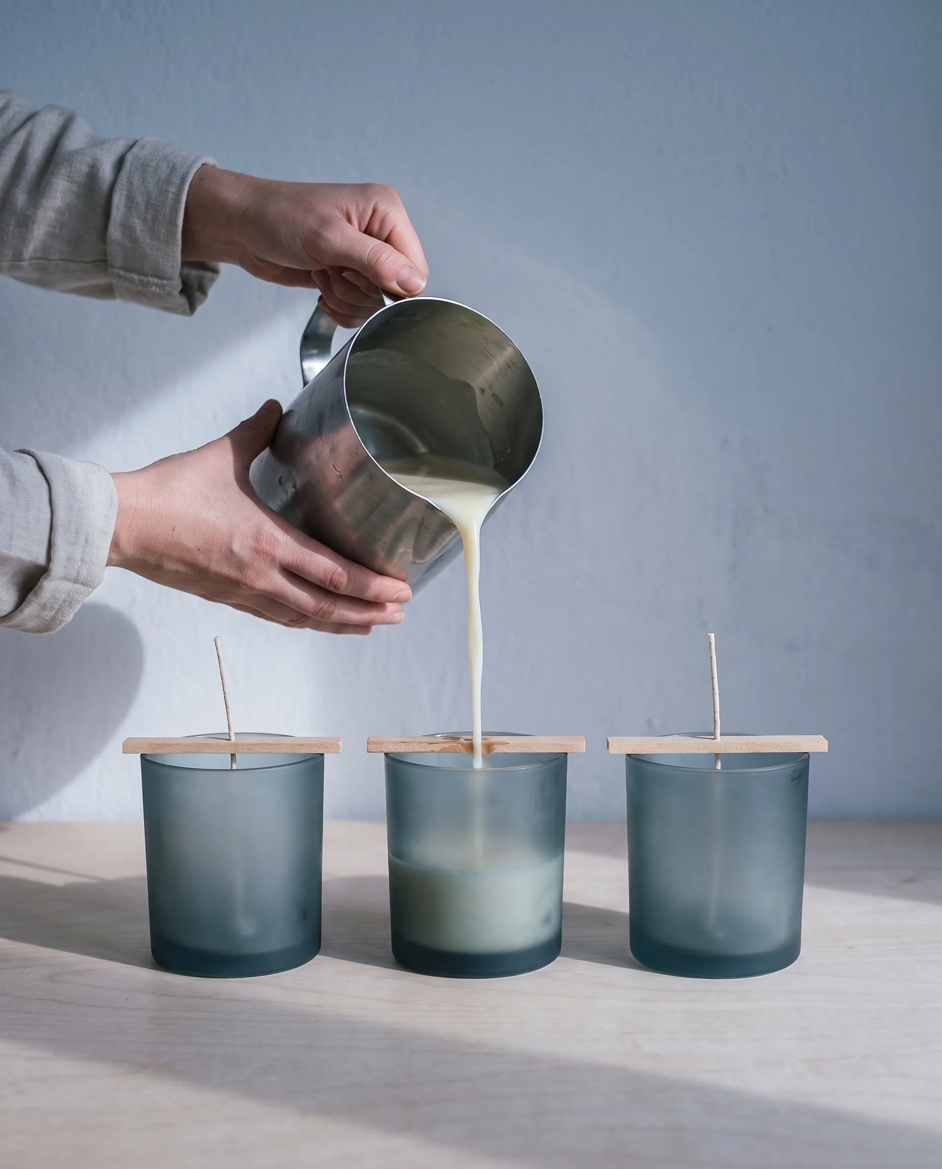 Hands pouring melted wax from a steel pitcher into a row of frosted glass tumblers on the bench.